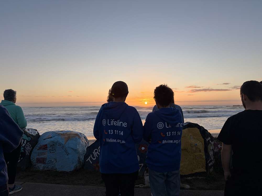 Walkers in Lifeline shirts standing at the rocks, watching the sun come up over the water